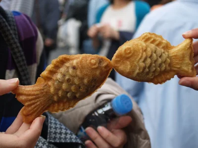 Kaksi henkilöä pitää ylhäällä taiyaki-leivoksia niin, että näyttää siltä kuin kalat suutelisivat