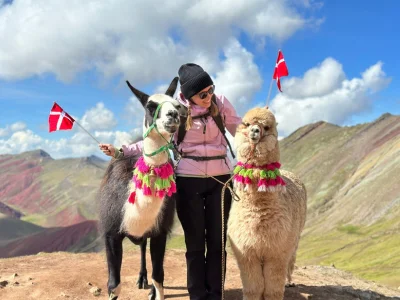 TourCompassin tiimin jäsen Iida seisoo kahden lipun ja alpakka vierellään Perun Rainbow Mountainin huipulla.